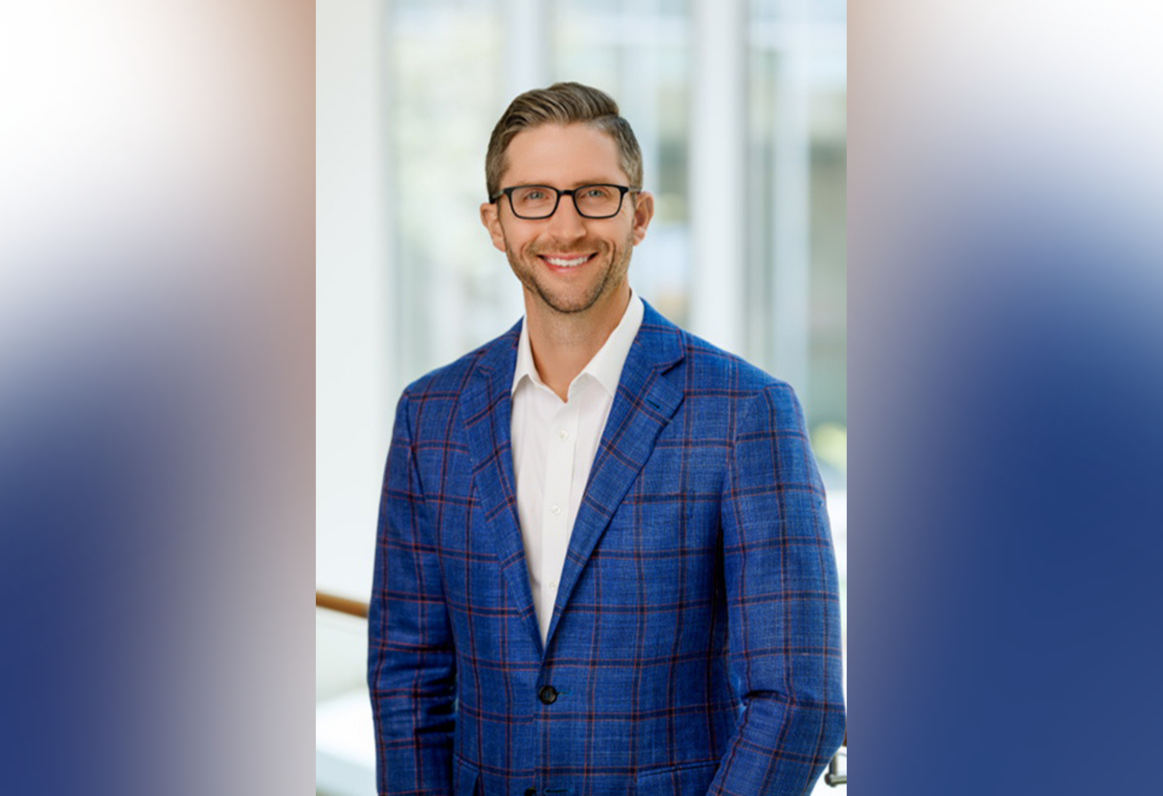 Portrait of Shafer. A smiling man in glasses and a blue checkered suit stands indoors. The background is softly blurred, suggesting a bright office or modern space.