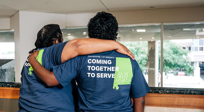 Two people in muted shirts with "UAB Medicine + St. Vincents, Coming Together to Serve AL" printed on the back, standing arm in arm, gazing out a window, conveying unity.