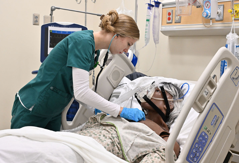 A student nurse in green scrubs uses a stethoscope on a patient simulator in a hospital bed. The patient wears an oxygen mask. Medical equipment surrounds them. The scene conveys care and attentiveness.