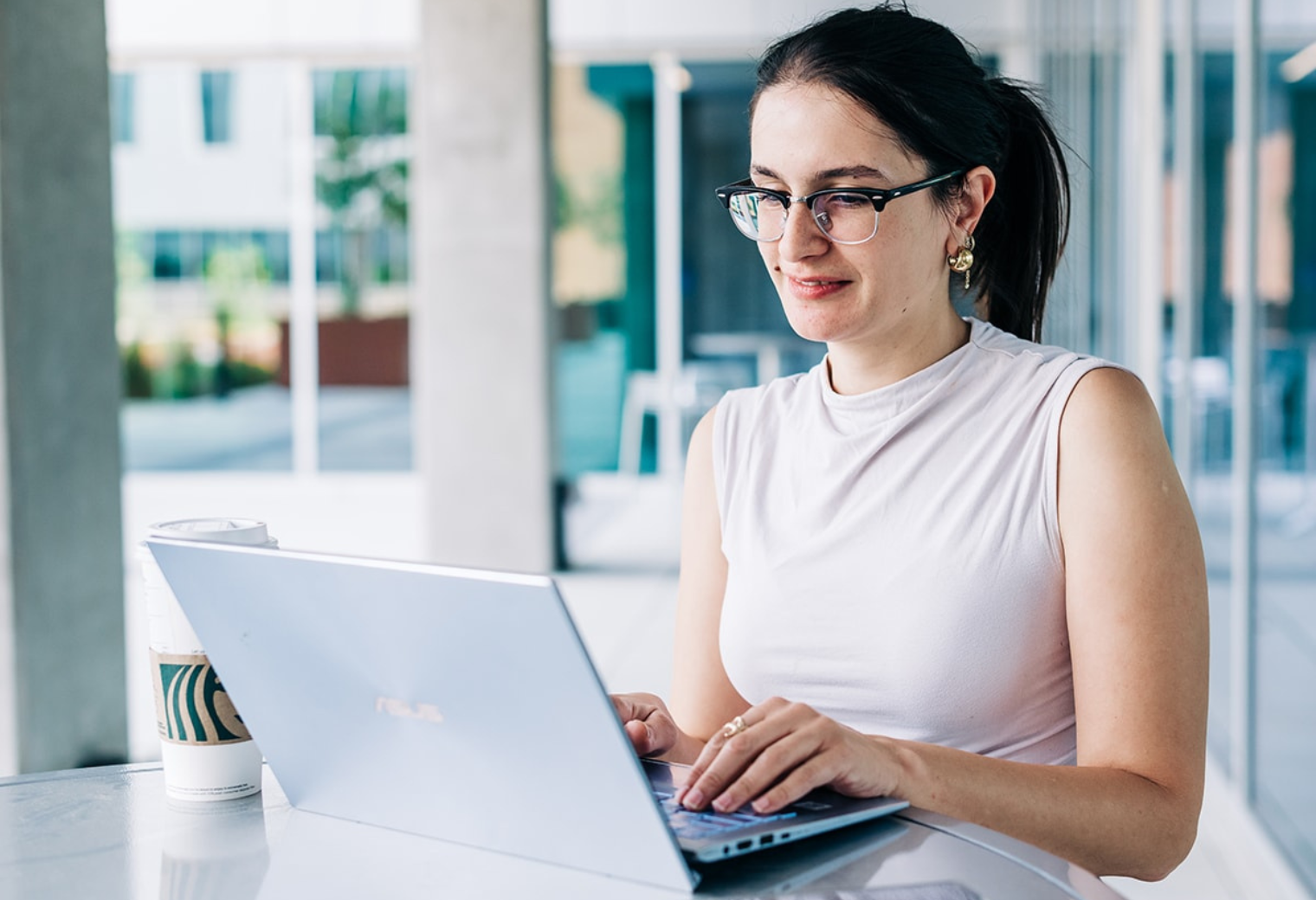 A woman with glasses and a ponytail is working intently on a laptop at a bright, modern café. A cup of coffee is beside her, reflecting focus and productivity.