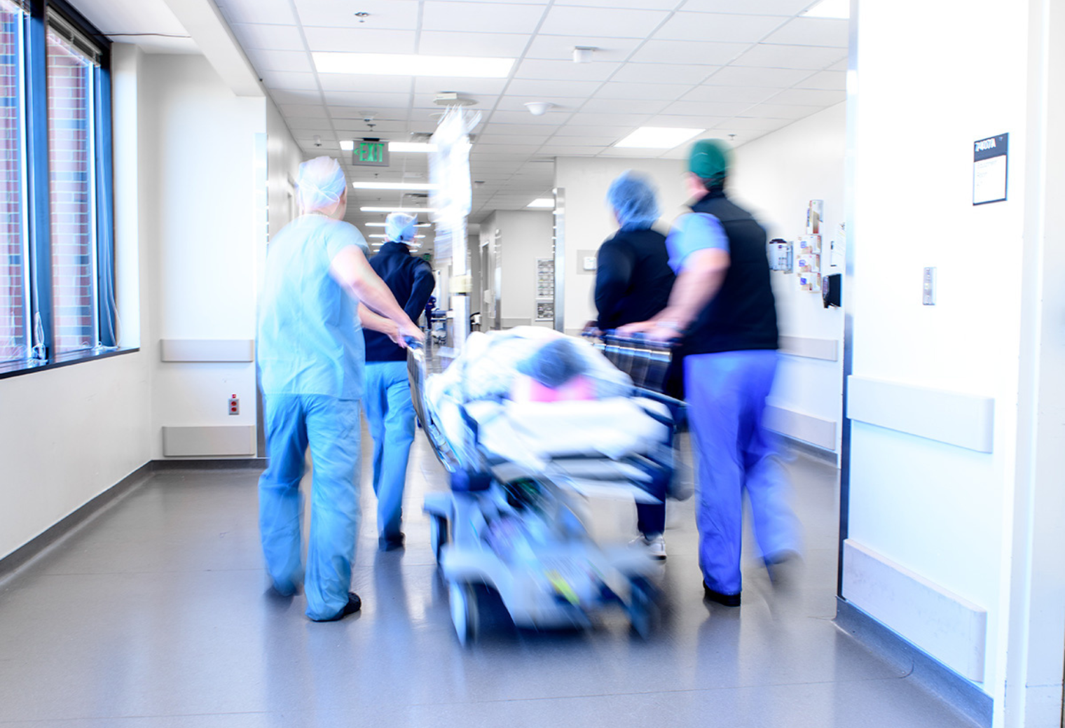 Medical team rushes a patient on a gurney through a bright hospital corridor, conveying urgency and efficiency. Blurred motion suggests haste.