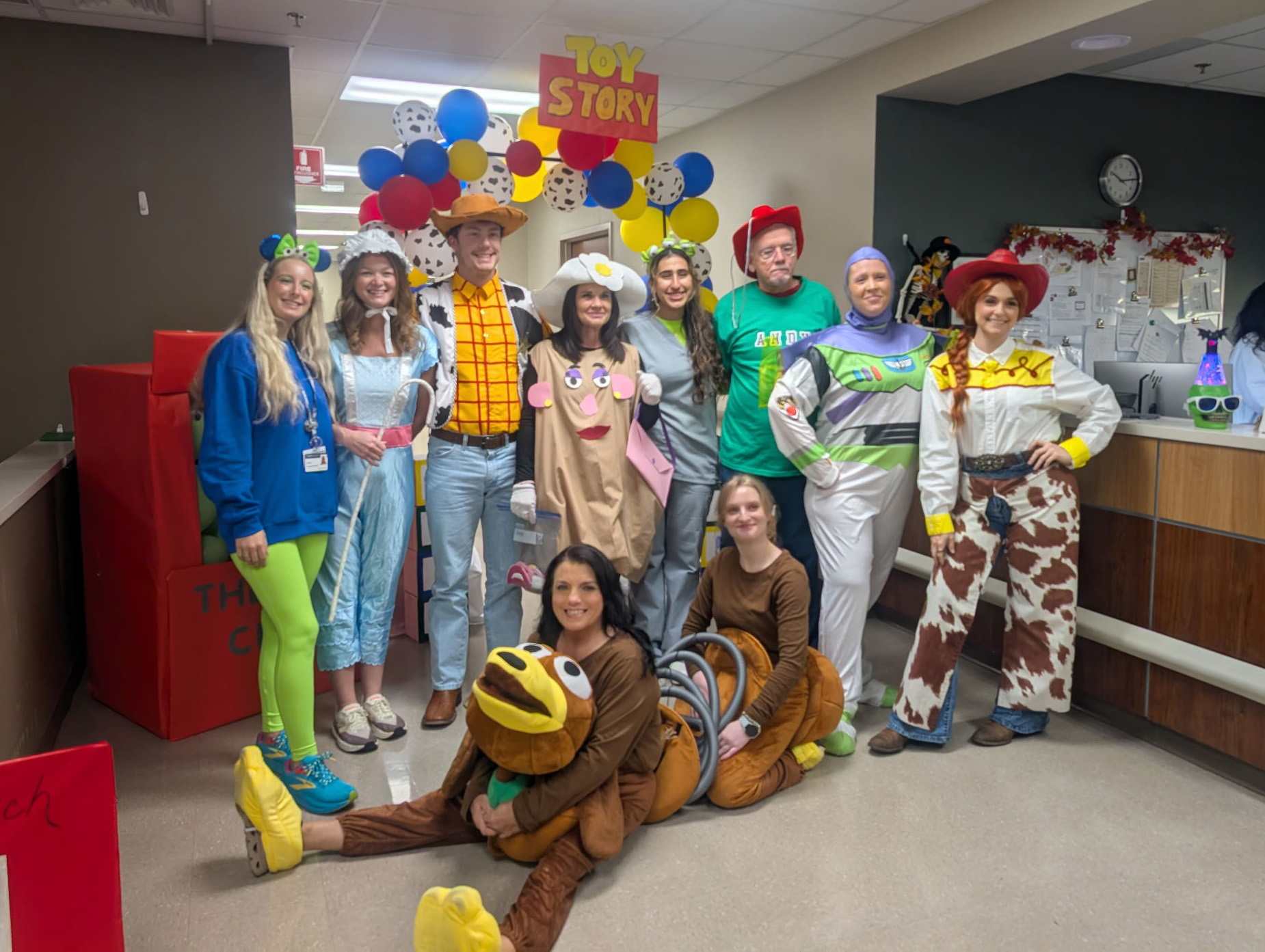 A group of people dressed in various "Toy Story" character costumes, smiling in a hallway. Colorful balloons and a "Toy Story" sign are in the background.