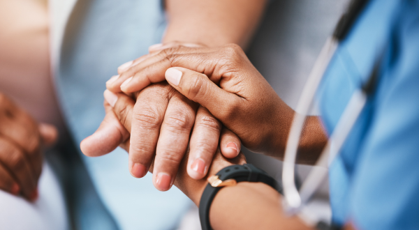 Two people gently holding hands, conveying support and care. One wears a blue uniform, suggesting a healthcare setting. The mood is comforting and empathetic.