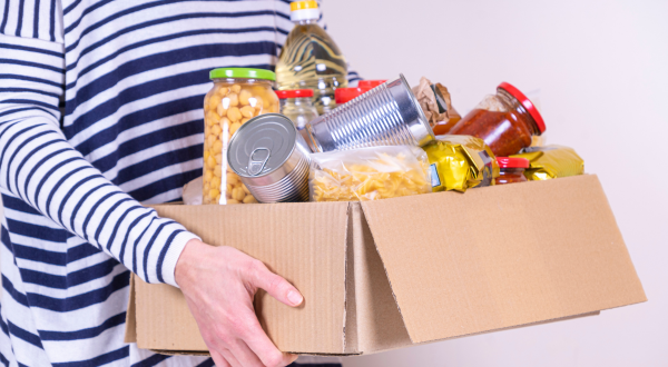 A person in a striped shirt holds a cardboard box filled with various groceries, including canned food, pasta, and jars. The scene is casual and organized.