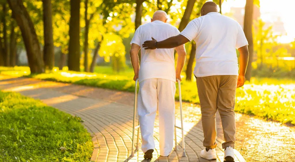 A caregiver gently assists an elderly man with a cane on a sunlit park path, surrounded by green trees, conveying support and compassion.