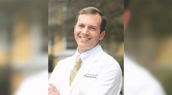Portrait of Dr. Colvard. Smiling man in a white lab coat with arms crossed, outdoors. He wears a yellow-striped tie. Background is a blurred mix of trees and buildings, conveying professionalism.