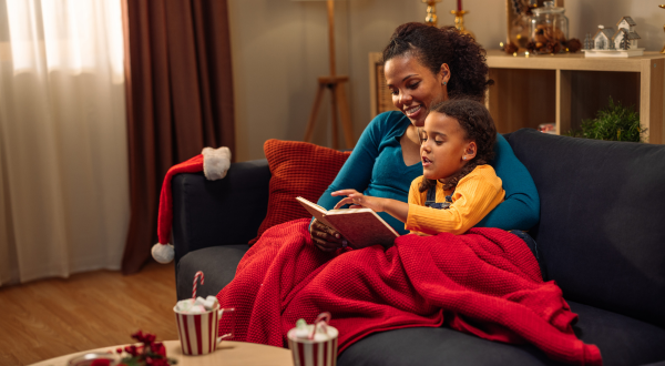 Mother reading a book to her daughter while they are sitting covered in blanket on the sofa during christmas holidays
