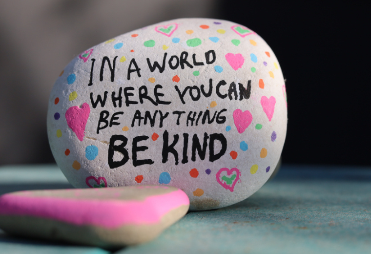 A painted rock with colorful dots and hearts reads "In a world where you can be anything, be kind." Another rock with pink edges is in the foreground.