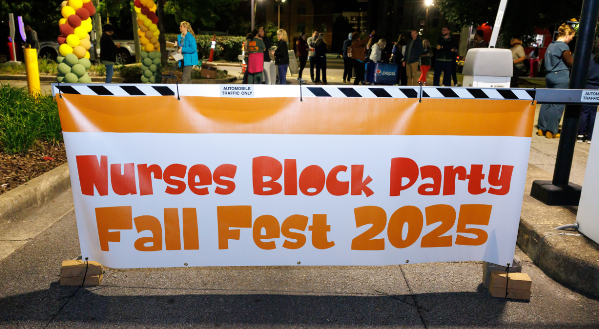 A vibrant banner with "Nurses Block Party Fall Fest 2025" in bold orange letters stretches across a street. Colorful balloons and a gathered crowd are visible in the background, creating a lively and festive atmosphere.