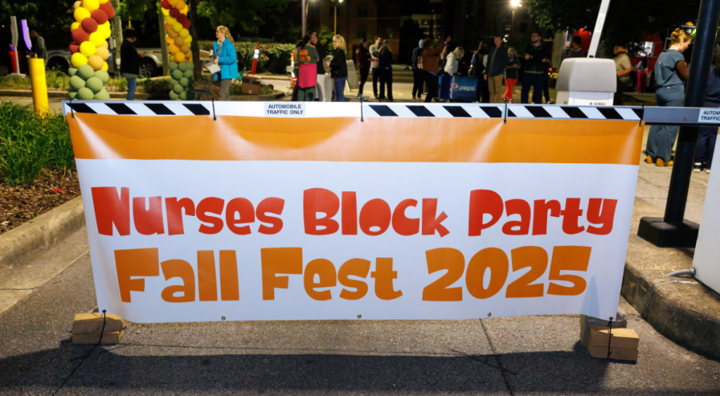 A vibrant banner with "Nurses Block Party Fall Fest 2025" in bold orange letters stretches across a street. Colorful balloons and a gathered crowd are visible in the background, creating a lively and festive atmosphere.