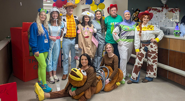 A group of people in colorful Toy Story costumes, including Woody, Buzz Lightyear, and Jessie, pose together indoors, smiling warmly. Festive atmosphere.