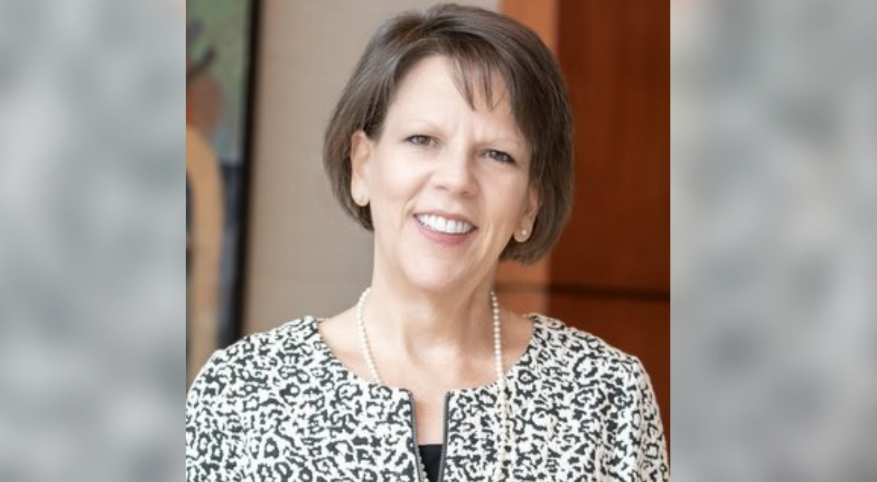 A smiling woman with short brown hair wears a black and white patterned jacket and a string of pearls. The background shows abstract wall art and wood paneling.
