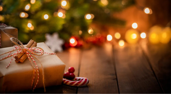A warmly lit Christmas scene with a wrapped gift tied with twine, cinnamon sticks, and a candy cane. Blurred tree and lights create a cozy, festive atmosphere.