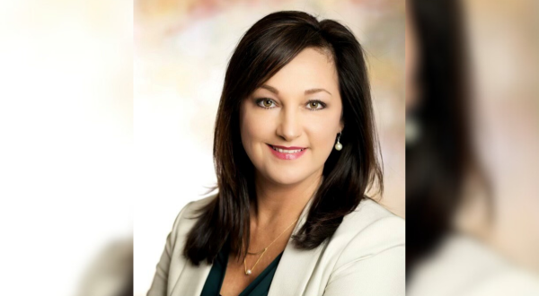 Portrait of Christy Nation. She is a woman with dark hair, smiling gently. She's wearing a light blazer, green top, earrings, and a necklace. Background is softly blurred.