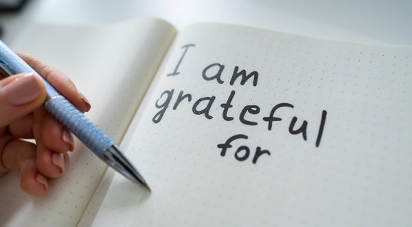 A hand holds a pen poised above an open notebook. The page features handwritten words, "I am grateful for," conveying a tone of reflection and thankfulness.