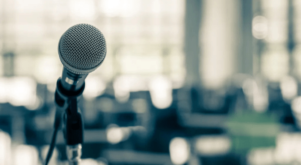Close-up of a microphone against a blurred, bright background suggesting an auditorium or conference hall. The scene conveys anticipation and focus.