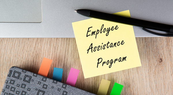 A sticky note reads "Employee Assistance Program" on a wooden desk next to a laptop. Various colorful tabs and a pen suggest organization and support.