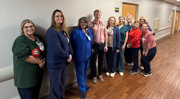 A group of nine healthcare workers, smiling, stand in a hospital corridor. They wear varied attire, creating a warm, welcoming atmosphere.