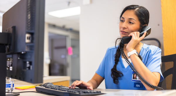 A nurse in blue scrubs sits at a desk, engaging on the phone while typing on a keyboard. Her expression is focused and attentive, setting a professional tone.