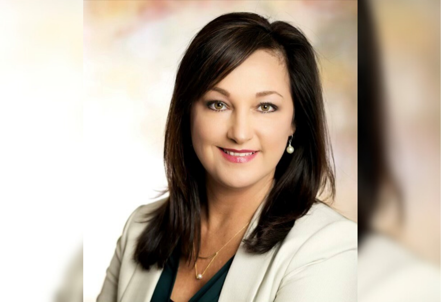Portrait of Christy Nation. She is a woman with dark hair, smiling gently. She's wearing a light blazer, green top, earrings, and a necklace. Background is softly blurred.