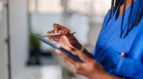 A person in blue scrubs uses a stylus on a tablet, suggesting a medical setting. The focus is on their hands, emphasizing technology in healthcare.