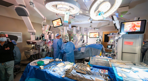 UAB robot kidney surgical team in blue scrubs operates in a high-tech operating room with robotic equipment. Monitors display images; trays of instruments are in the foreground.