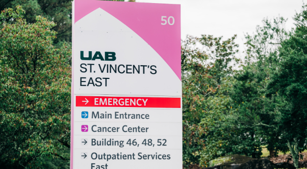 A hospital sign for UAB St. Vincent's East lists directions to the Emergency Room, Main Entrance, Cancer Center, and other facilities, amidst lush greenery.