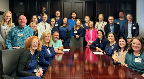 A group of smiling medical professionals and staff pose around a large wooden conference table. The mood is friendly and collaborative.