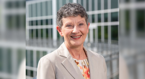 Portrait of Terri Poe. A woman with short gray hair smiles warmly, wearing a beige blazer over a floral top. She stands outdoors, with a modern glass building in the background.