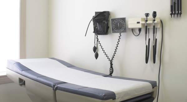 An empty medical examination room with a padded table covered in white paper, wall-mounted blood pressure monitor, and otoscope. The setting is clean and clinical.