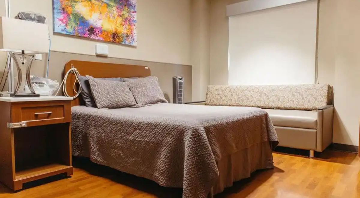 A hospital room with a cozy, calm vibe. It has a neatly-made bed with gray bedding, a small side table, a patterned couch, and a colorful abstract painting on the wall.