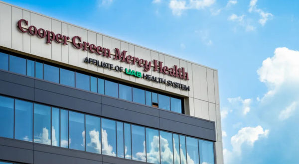 A modern building exterior with "Cooper Green Mercy Health, Affiliate of UAB Health System" on top. The blue sky with clouds reflects in large windows.