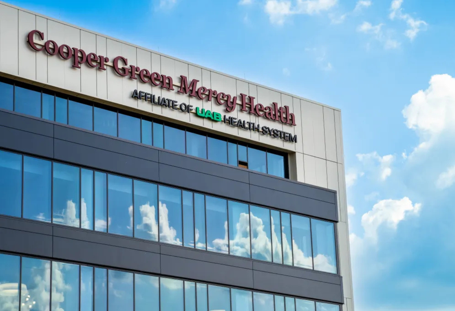 A modern building exterior with "Cooper Green Mercy Health, Affiliate of UAB Health System" on top. The blue sky with clouds reflects in large windows.