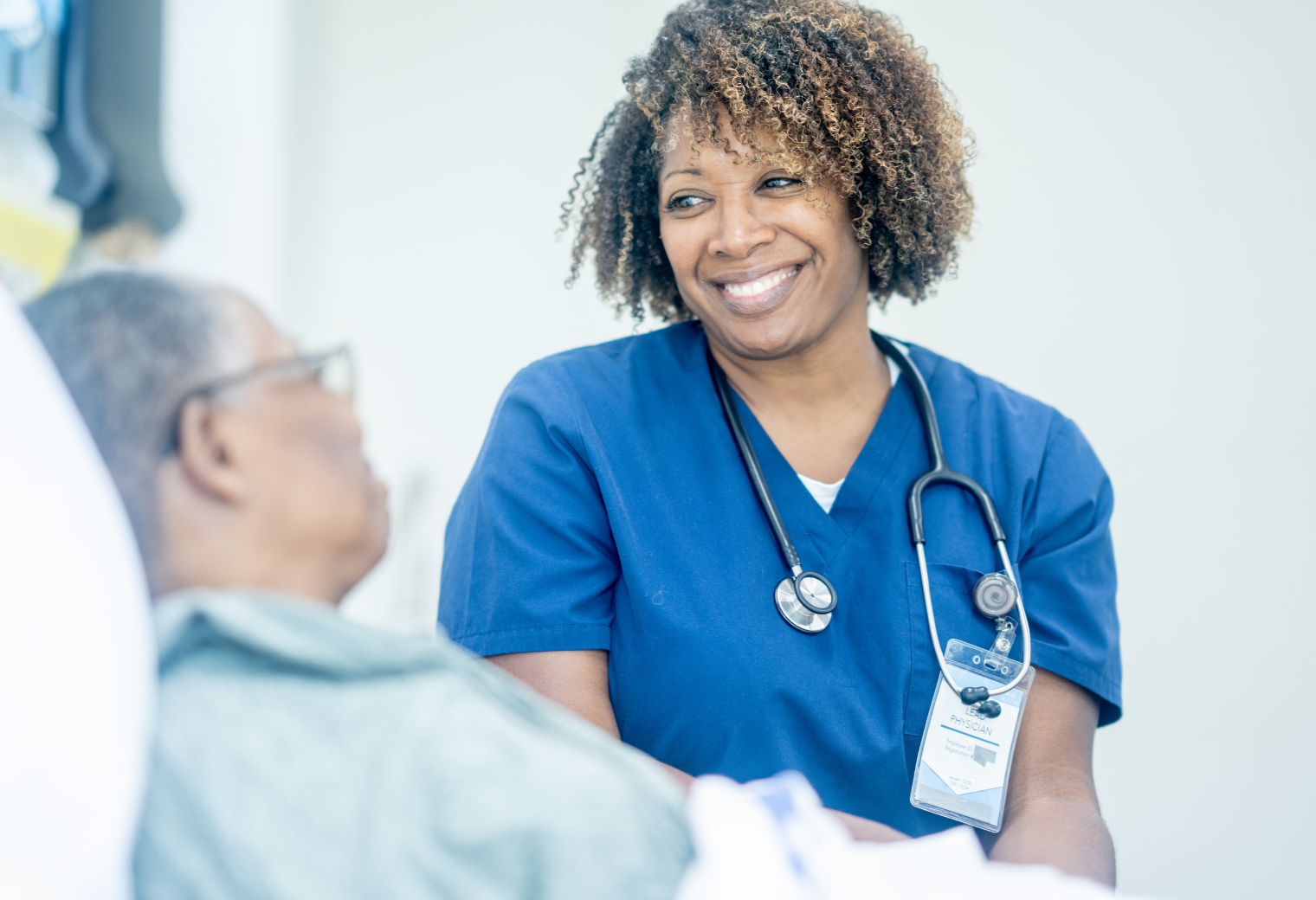 A nurse in blue scrubs with a stethoscope smiles warmly at a patient in a hospital setting. The atmosphere is caring and professional.