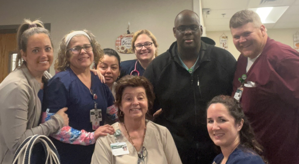 Robin Wadsworth surrounded by coworkers. A group of eight smiling medical professionals, in scrubs and ID badges, pose together in a hospital setting, conveying a sense of teamwork and camaraderie.