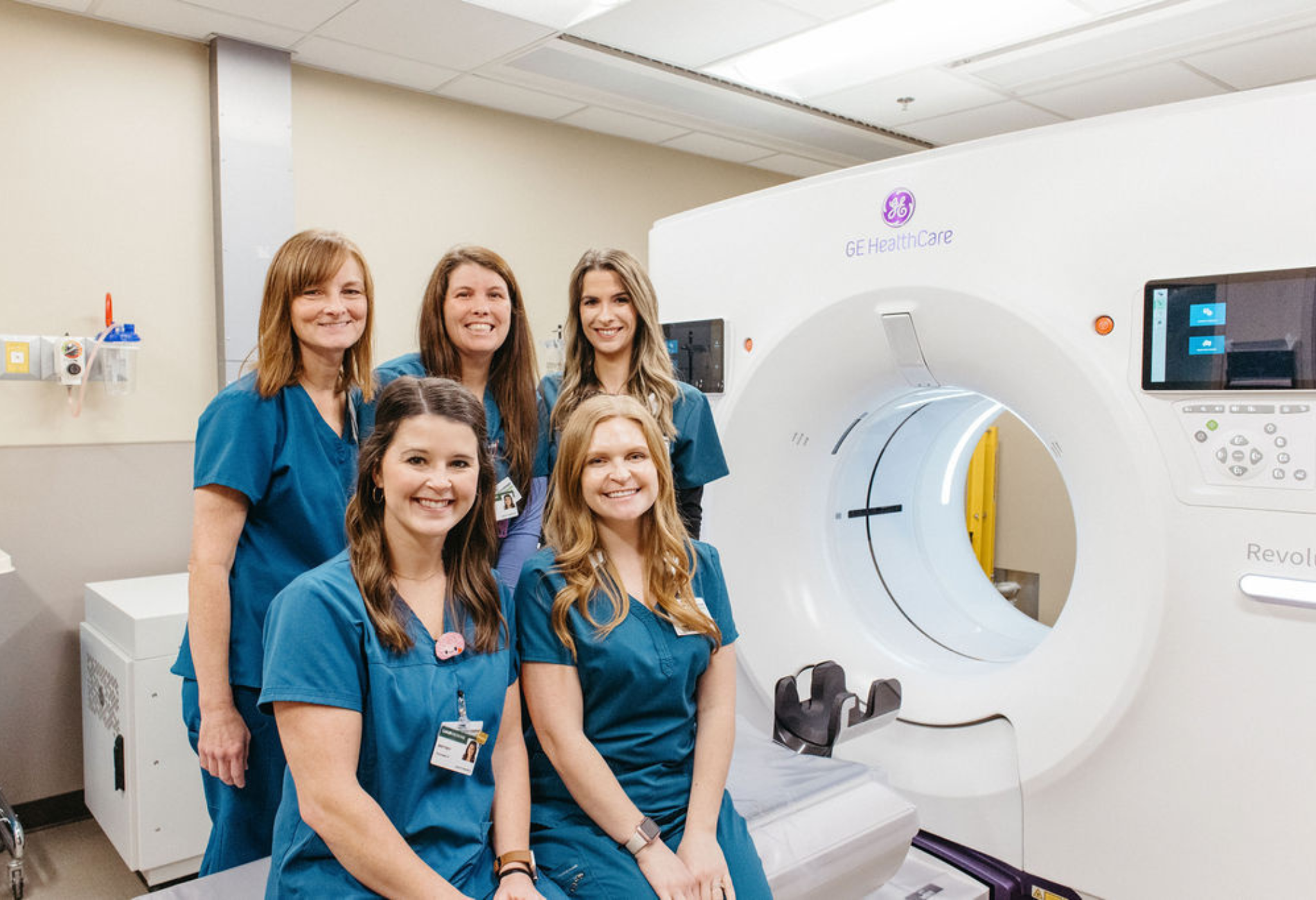 Five medical professionals in blue scrubs smile beside a large, white CT scanner in a hospital room, conveying a sense of teamwork and professionalism.
