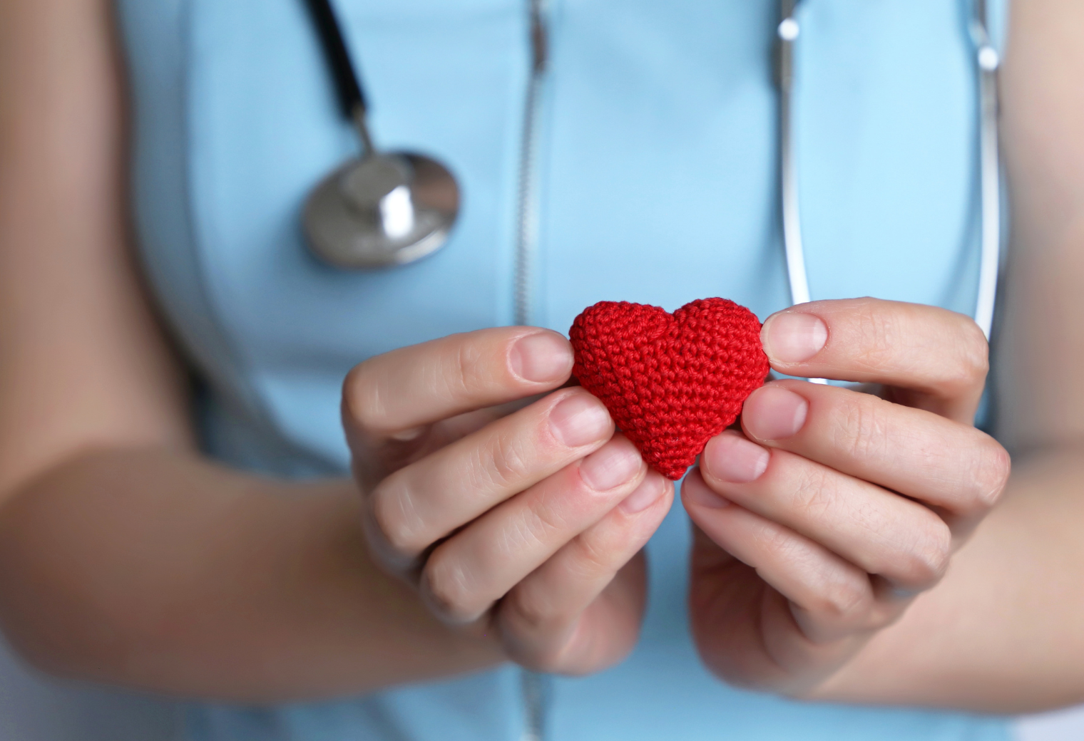 Hands of a person in a blue medical uniform gently holding a small, knitted red heart. A stethoscope hangs around their neck, suggesting care and compassion.