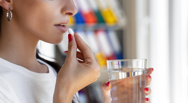 Close-up of a woman holding a pill and a glass of water, about to take the pill. Her expression is focused and calm. Background shows blurred shelves.