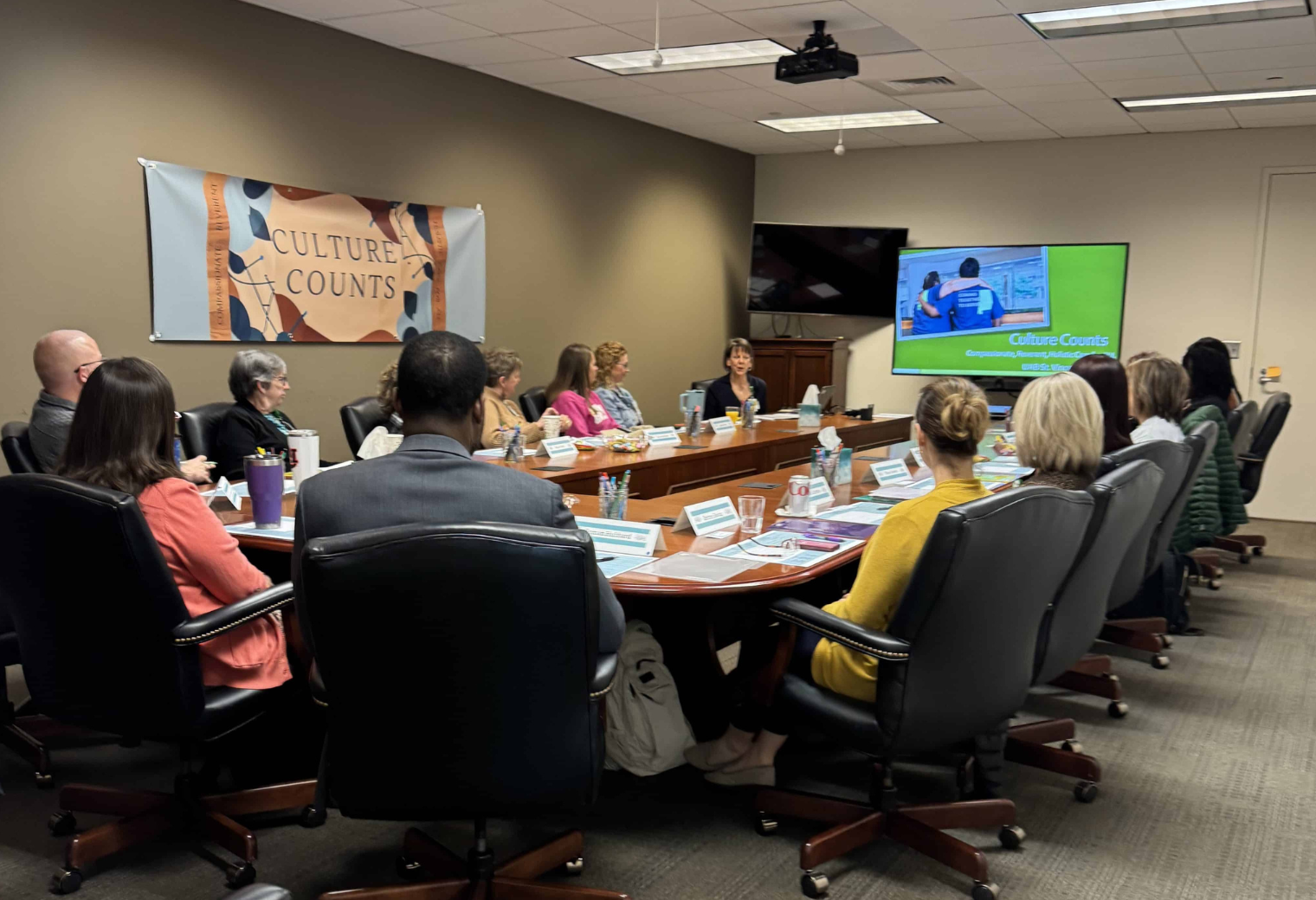 A group of people sitting around a large wooden U-shaped conference table during a "Culture Counts" meeting. A television screen in the background shows a photo of a person experiencing homelessness.