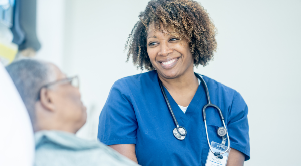 A nurse in blue scrubs with a stethoscope smiles warmly at a patient in a hospital setting. The atmosphere is caring and professional.