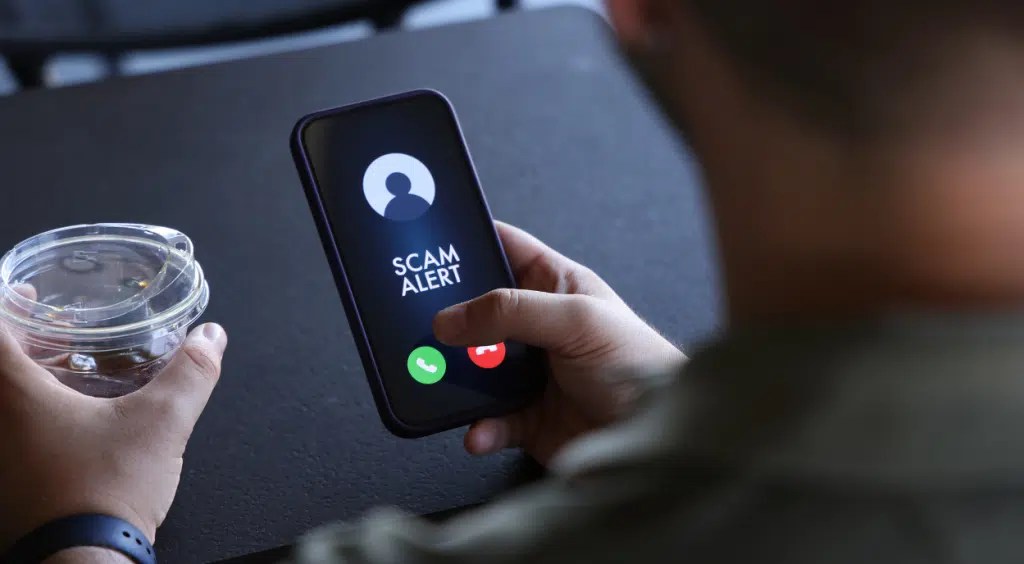 Man holding a smartphone showing a "Scam Alert" call on screen, with red and green buttons. He holds a plastic cup lid in the other hand. Alert, technology.
