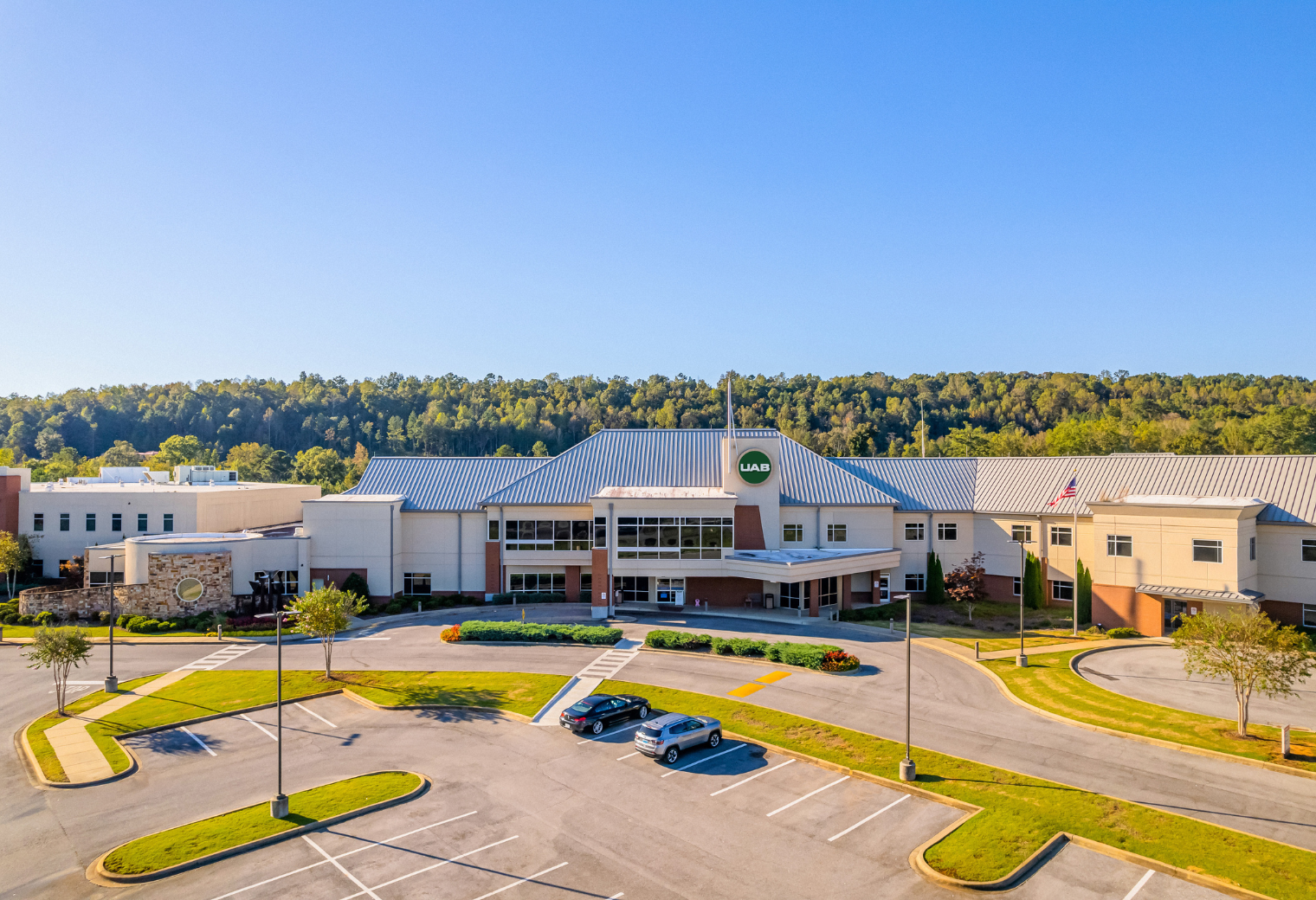 Outside photo of UAB Medicine St. Clair. Aerial view of a modern medical building with a UAB sign, set against a backdrop of lush greenery and clear blue sky, conveying a serene atmosphere.
