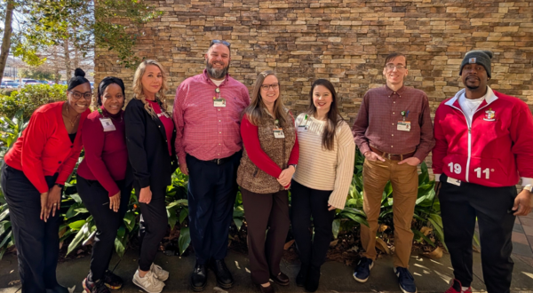 A group of eight people stand smiling in front of a stone wall. Most are wearing red or maroon, conveying a cheerful and friendly atmosphere.