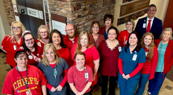 A group of 18 healthcare workers, mostly women, pose smiling in a hospital corridor. Many wear red, conveying unity and support.