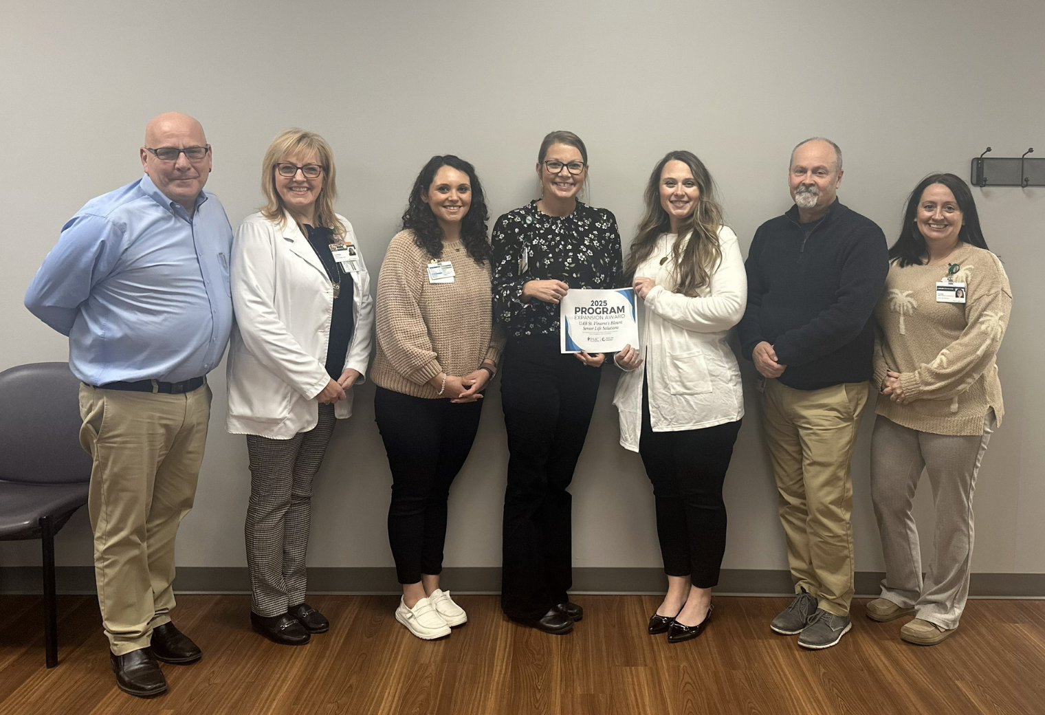 A group of seven people, smiling, stand together indoors against a beige wall. One person holds a certificate. They appear happy and professionally dressed.