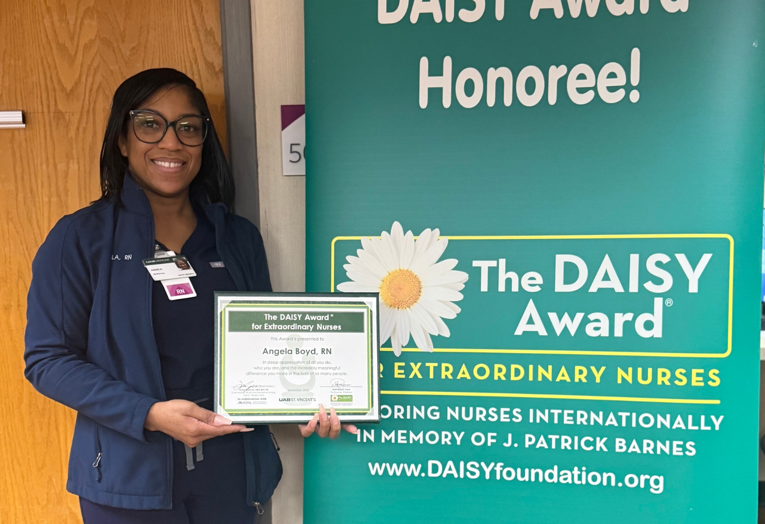 Angela Boyd, RN, smiling and holding her framed DAISY Award for Extraordinary Nurses while standing next to a large green DAISY Foundation banner.