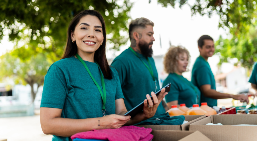A smiling woman in a teal volunteer shirt holds a tablet while standing with a group of volunteers at an outdoor community service event.