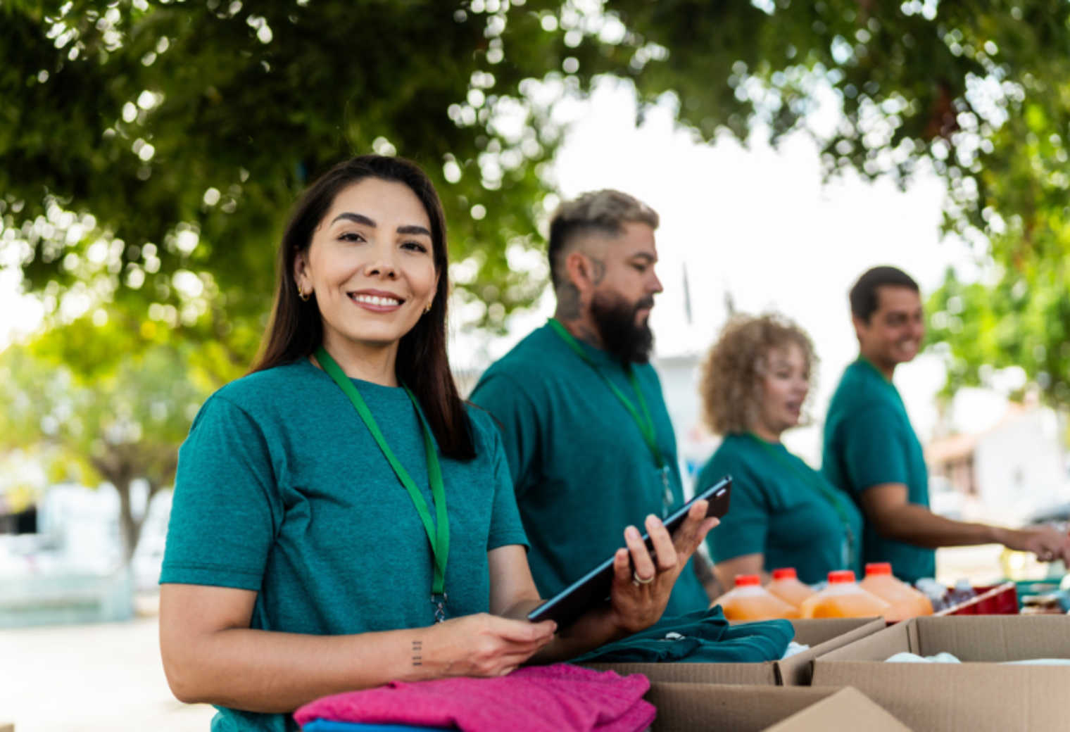 A smiling woman in a teal volunteer shirt holds a tablet while standing with a group of volunteers at an outdoor community service event.