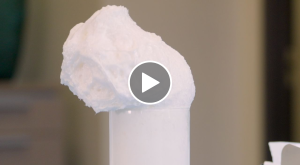A close-up video thumbnail showing a large, textured mound of white chemical foam overflowing from a clear glass cylinder, with a play button icon centered on the image.