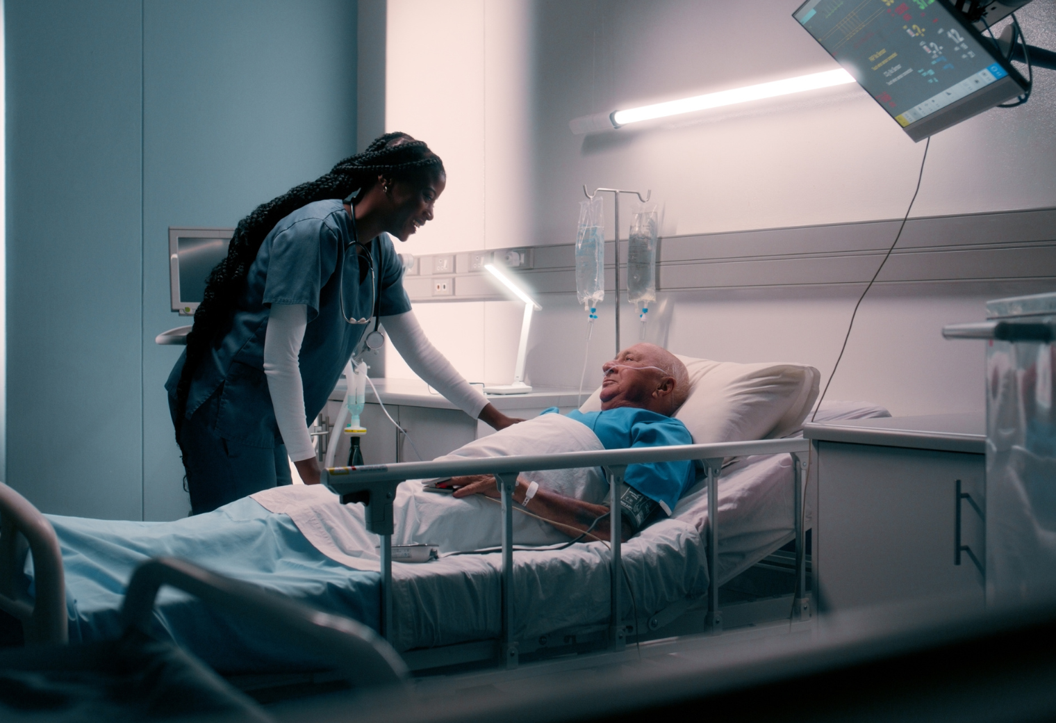 A female nurse in teal scrubs provides bedside care to an elderly male patient in a hospital room.